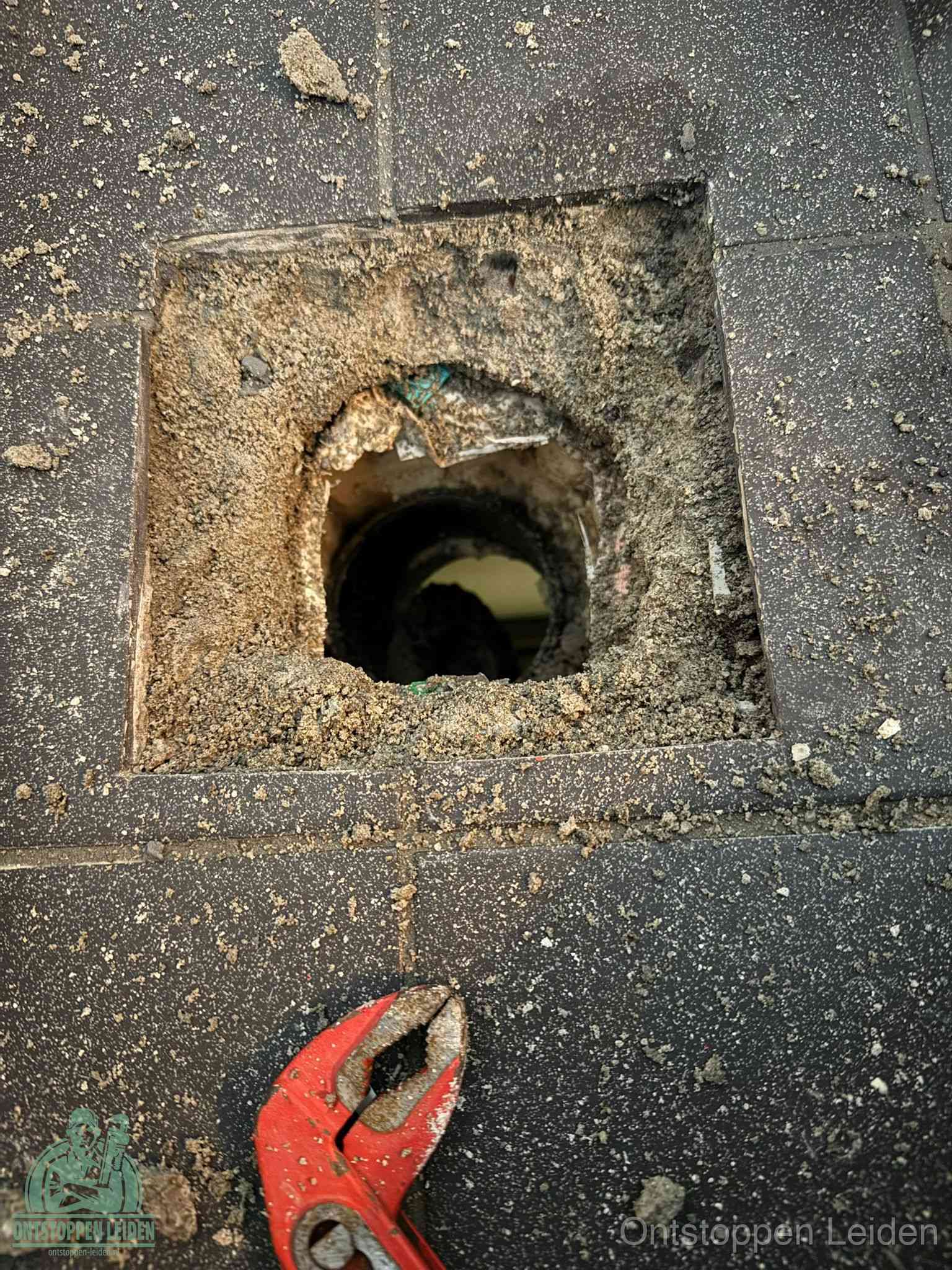 Een geopend rioolputje in een betontegelvloer met losgemaakt rooster en een zichtbare ronde afvoerbuis, het achterhalen waar de afvoerverstopping zit in Leiden