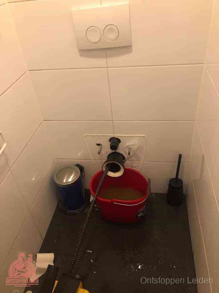 Een ontstopte wand-wc met verwijderde toiletpot waar een ontstoppingsveer in de wanduitgang verdwijnt, het achterhalen waar de verstopping zit in Leiden