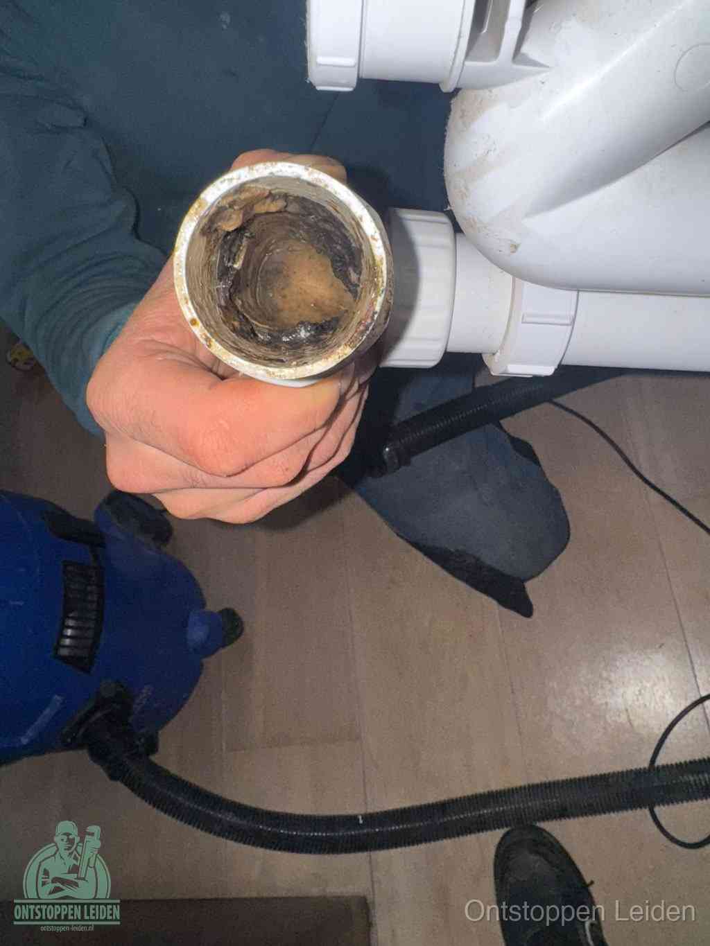 Een hand houdt een losgekoppeld PVC-afvoerbuisstuk vast dat van binnen volledig is dichtgeslibd met vet, vuil en aanslag, de meest voorkomende oorzaak van een verstopte afvoer in Leiden