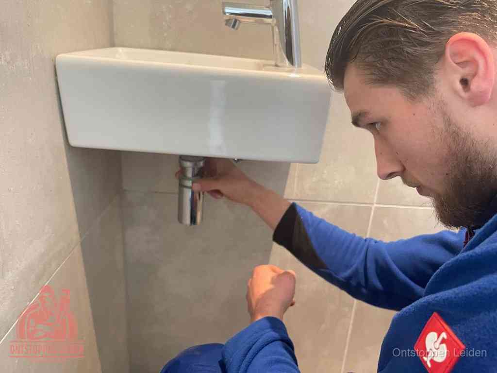 Een monteur draait met de hand de zwanenhalssifon onder een fonteinwasbak in de toiletruimte los om de bron van rioollucht in Leiden te vinden