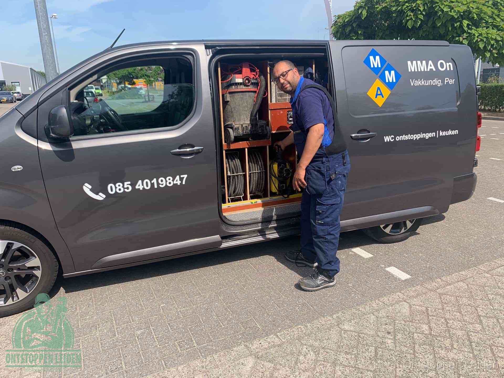 Een monteur bij de open schuifdeur van zijn bedrijfsbus op een parkeerplaats, klaar om de oorzaak van terugkerende afvoerverstoppingen aan te pakken in Leiden